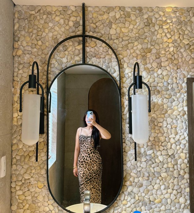 A woman in a leopard-print dress takes a mirror selfie in an upscale bathroom featuring a stone pebble wall, oval mirror, and elegant pendant sconces, capturing a stylish "good morning" moment before diving into her busy content-creator schedule.