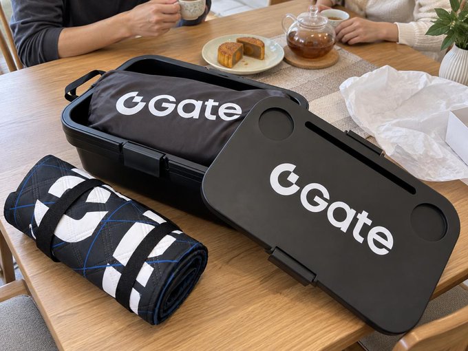 A black camping picnic kit branded "Gate" is displayed on a wooden table, featuring a folded insulated bag, a rolled-up plaid mat, and a hard case—all items referenced in the post as gifts from @Gate that humorously prove the author can no longer use the excuse of lacking gear for camping.