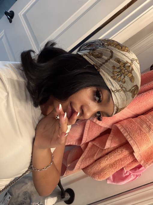 A woman wearing a white outfit and a patterned silk headscarf poses with her hands near her face, with a pink towel in the background.