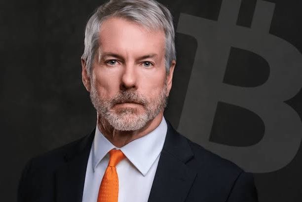 A distinguished man with gray hair and a beard, wearing a dark suit with an orange tie, stands confidently in front of a large Bitcoin (₿) symbol. The image likely represents Michael Saylor, the executive chairman of Strategy (formerly MicroStrategy), whose company has returned to profitability as Bitcoin surpasses $75,000.