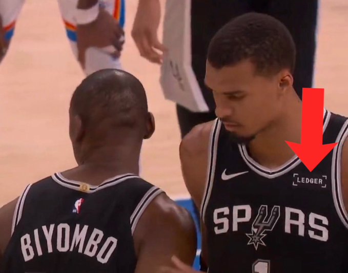 The image shows a basketball player wearing a black and white jersey with the name "Lerner" written on his back, standing next to another player who has a red arrow pointing at him. The background of the photo reveals that they are inside a sports arena or court setting.