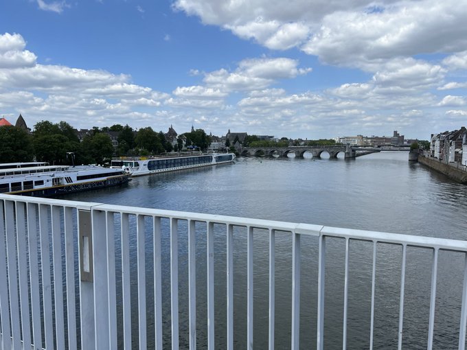View from a bridge over a river with a stone arch bridge, boats docked along the bank, and a city skyline in the distance under a cloudy sky.