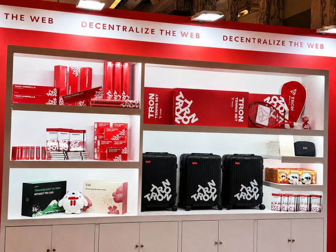 A TRON-branded merchandise display with the slogan "THE WEB DECENTRALIZE THE WEB" featuring red umbrellas, tennis sets, books, black suitcases, and other promotional items on white shelves.