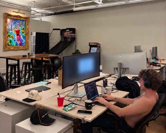 A shirtless person wearing headphones works on a laptop at a desk in a modern office space featuring dual monitors, a basketball arcade game, and an Elmo painting.