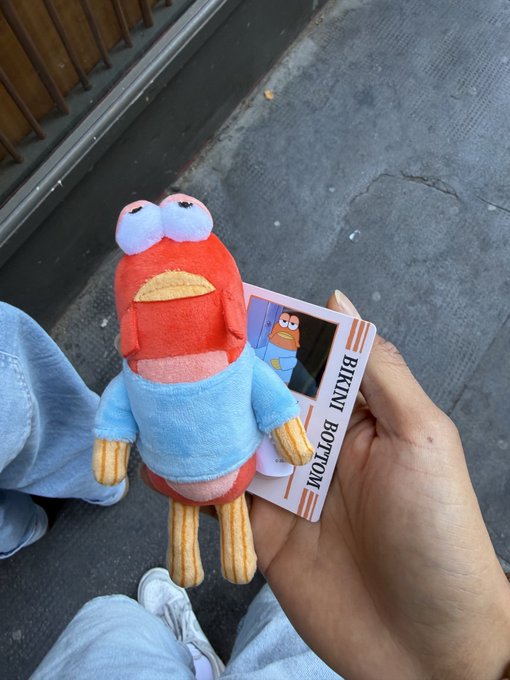 A hand holds up a small plush toy of Bikini Bottom's character (with a tag reading "BIKINI BOTTOM") along with a card displaying the cartoon character, while the person wears light blue jeans and stands on a sidewalk. The image ties into the post's playful nod to "Bob" by featuring a recognizable Bikini Bottom character plush, likely meant to bring attention to a Bob-themed meme or crypto token.