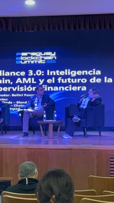 Two men seated in armchairs on a stage at the 'Paraguay Blockchain Summit,' discussing 'Compliance 3.0: Inteligencia [artificial], AML y el futuro de la supervisión financiera' with moderator Bellini Foss and another panelist.