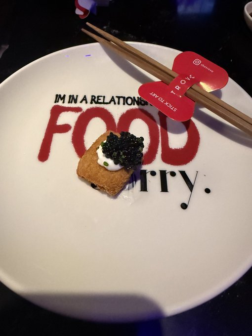 A small gourmet appetizer topped with black caviar and cream served on a white plate that reads 'I'M IN A RELATIONSHIP WITH FOOD', accompanied by wooden chopsticks on a red rest.