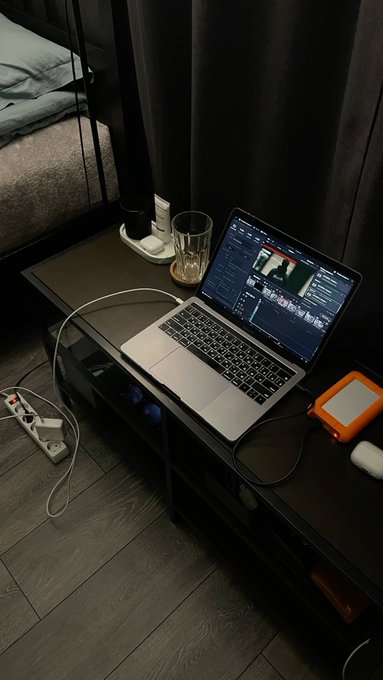 A laptop displaying video editing software sits on a dark desk in a dimly lit room, accompanied by a glass, cables, and a small orange device.