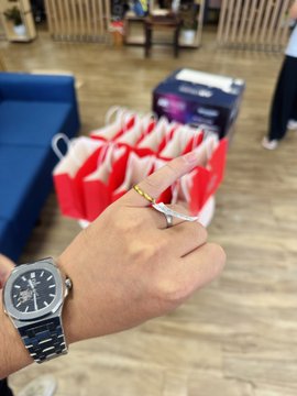 A first-person view of a hand wearing a silver wristwatch and a gold ring, holding a pen, with red and white gift bags visible in the background on a wooden floor.