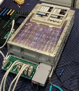 A restored Apollo Guidance Computer (AGC) is shown operating on a blue workbench, with its exposed circuitry, wiring harnesses, and external interfaces visible, illustrating the vintage hardware that Ken Shirriff reportedly programmed to perform bitcoin mining hash calculations.