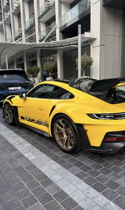 A bright yellow Porsche 911 GT3 RS parked on a brick driveway, showing its large rear wing and bronze-colored wheels.