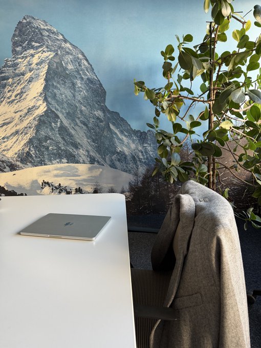 A MacBook rests on a white desk with a gray coat draped over a chair, framed by a stunning backdrop of a snow-capped Swiss mountain (likely the Matterhorn) and lush greenery—capturing the perfect balance of focused remote work and breathtaking alpine scenery that defines "Swiss office days."