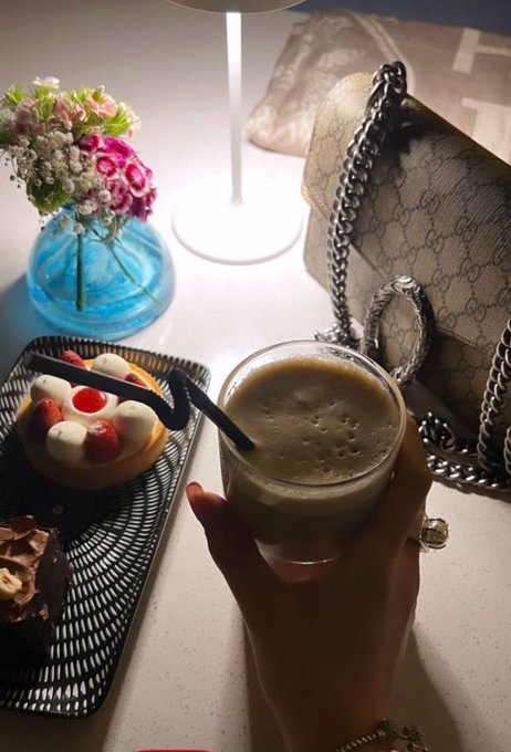 A hand holds a glass of iced coffee next to a designer handbag, a vase of pink flowers, and a plate of fruit-topped pastries and desserts.