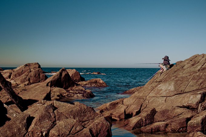 A scenic view of a person fishing from rocky cliffs overlooking a vast, deep blue ocean under a clear sky.