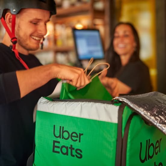 A smiling Uber Eats delivery driver wearing a helmet holds a green insulated delivery bag while receiving an order from a cashier in a restaurant.