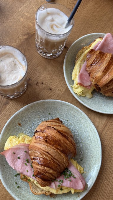 An overhead photo of two plates with ham, scrambled eggs, and croissants on a wooden table, accompanied by two glasses of iced coffee or latte.