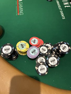 A collection of WSOP (World Series of Poker) chips in various denominations ($5,000, $1,000, $100) and colors are stacked on a green felt poker table, with text visible reading "SIDE BET EVENTS" and "GAMING" on the table surface. This relates to the post about being "still alive" in the Gladiators Poker Tournament at the WSOP, showing the chips in play during the competition.