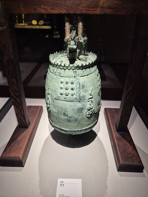 An ancient bronze 'Bell' artifact with green patina displayed in a museum case, featuring decorative figures on top and raised knobs on its surface.