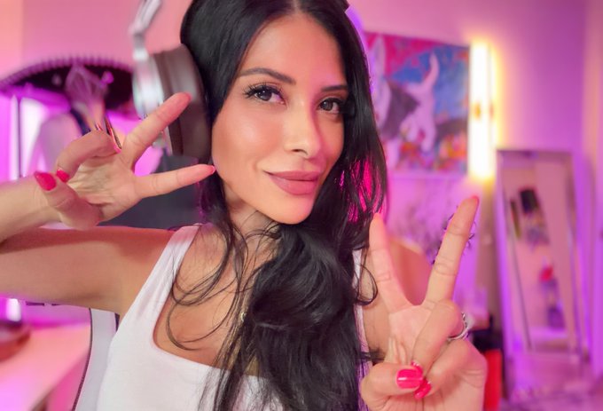 A woman with long dark hair wearing headphones poses with peace signs, smiling at the camera in a room lit with pink and purple neon lighting.