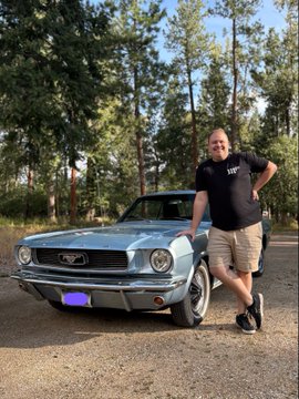 A smiling man in a black t-shirt and tan shorts leaning against a classic light blue Ford Mustang parked on a dirt road with a forest in the background.