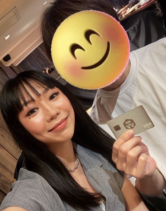 A selfie of a smiling woman holding up a Visa card, with a person's face obscured by a large smiling emoji sticker.