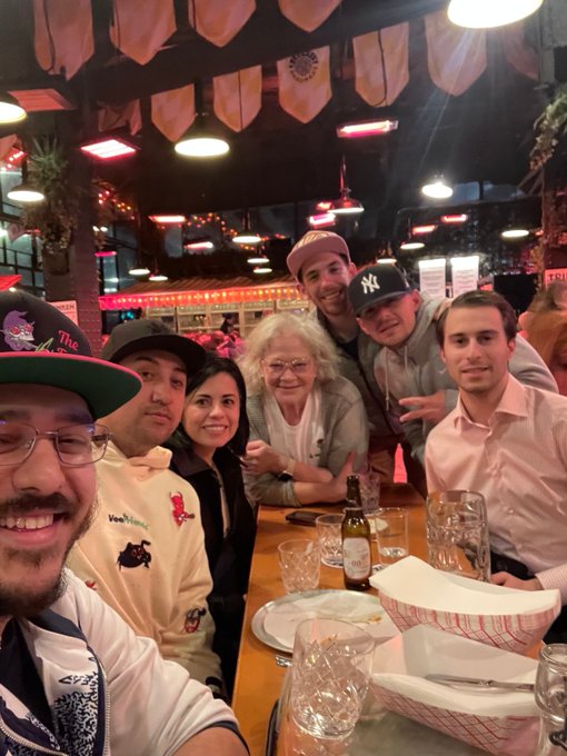 A group of six people pose for a selfie at a restaurant table, including an older woman with curly blonde hair and several younger adults wearing baseball caps, under string lights and decorative pennants.