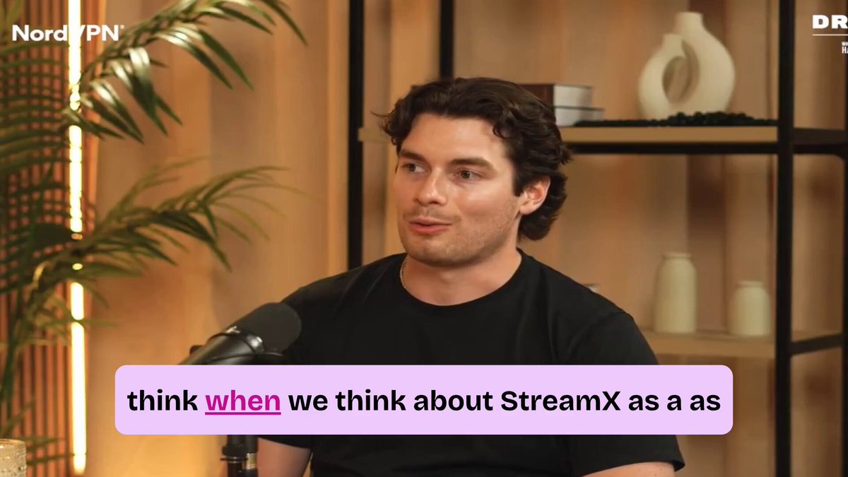 The image features a man wearing a black shirt sitting at a desk with a microphone in front of him. The background includes a plant and a shelf holding various items, such as vases and books. There are also some text elements in the image that read "Focus on what is in the image and how it relates to the post".