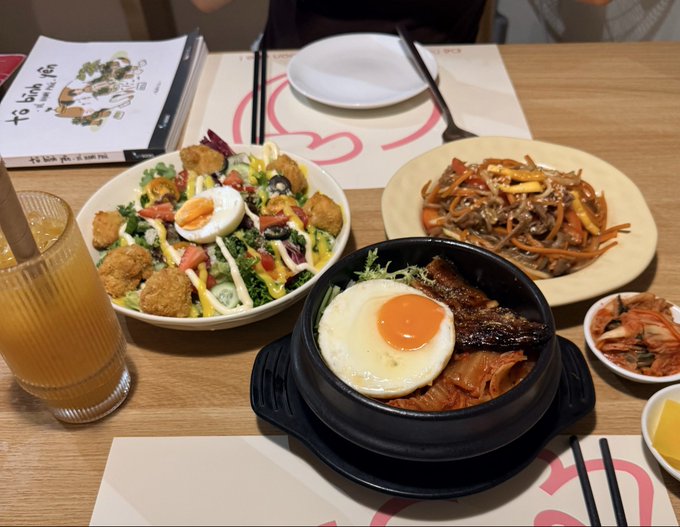 A Korean restaurant meal on a wooden table, featuring bibimbap with a fried egg, a salad with chicken nuggets, japchae, kimchi, and a glass of iced tea.