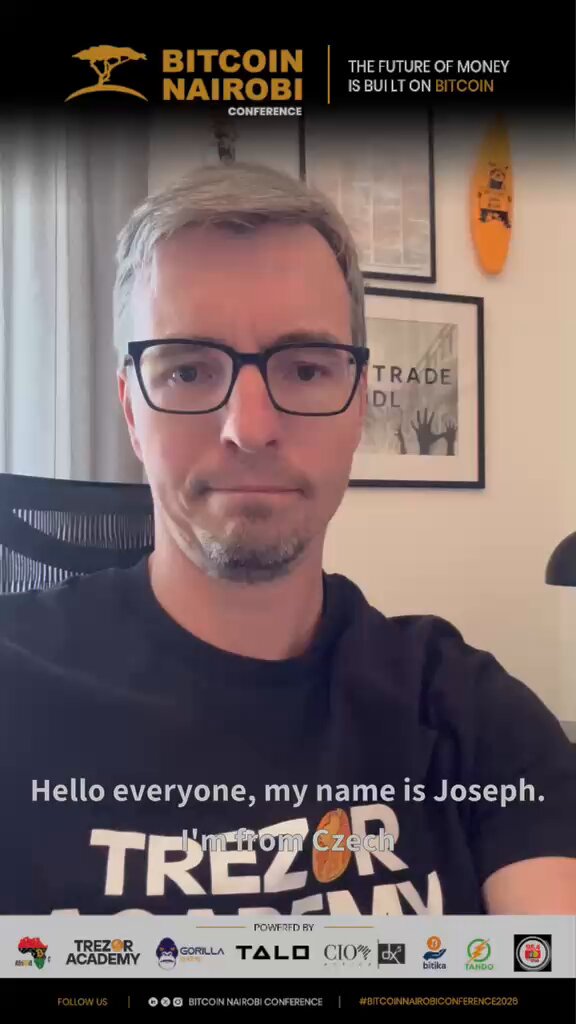 The image features a man wearing glasses, sitting at a desk with his arms crossed. He has a black t-shirt that says "Hello everyone, my name is Joseph." The background of the image includes text about an upcoming Bitcoin Nairobi conference, along with logos for other companies and organizations involved in the event.