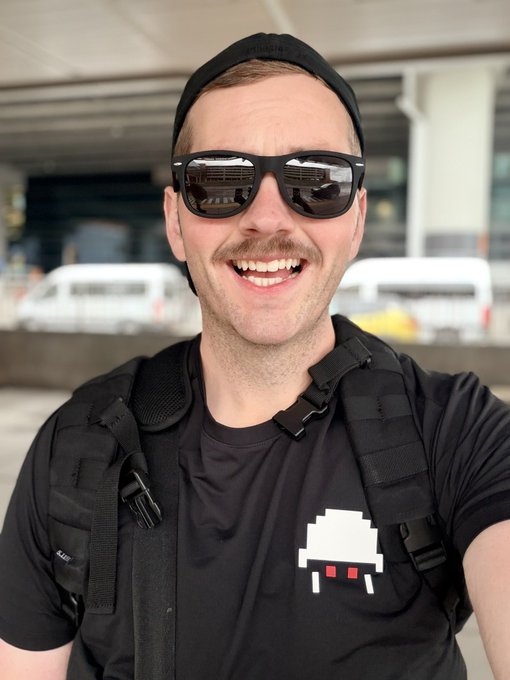 A smiling man wearing sunglasses and a backward cap takes a selfie while wearing a black backpack and a t-shirt featuring a pixel-art space invader design.