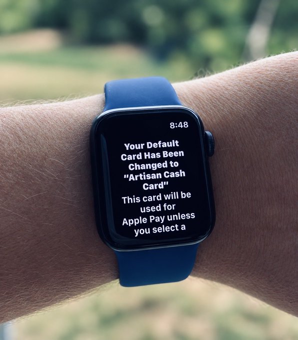 A wrist wearing a blue Apple Watch displays a notification at 8:48 reading "Your Default Card Has Been Changed to 'Artisan Cash Card'. This card will be used for Apple Pay unless you select a[nother]" — capturing a tech moment that contrasts with the post's "Touching grass" message about disconnecting from devices outdoors.