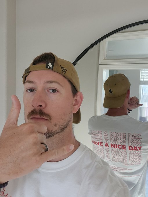 A selfie of a man with a mustache wearing a backwards tan cap, making a hand gesture, with a mirror reflection showing the back of his white t-shirt printed with 'HAVE A NICE DAY'.