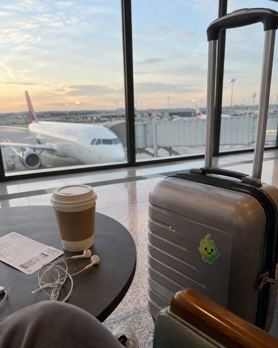 A travel scene at an airport gate shows a coffee cup, earbuds, and a boarding pass on a table next to a suitcase with a green flame sticker. An airplane is visible through the window.