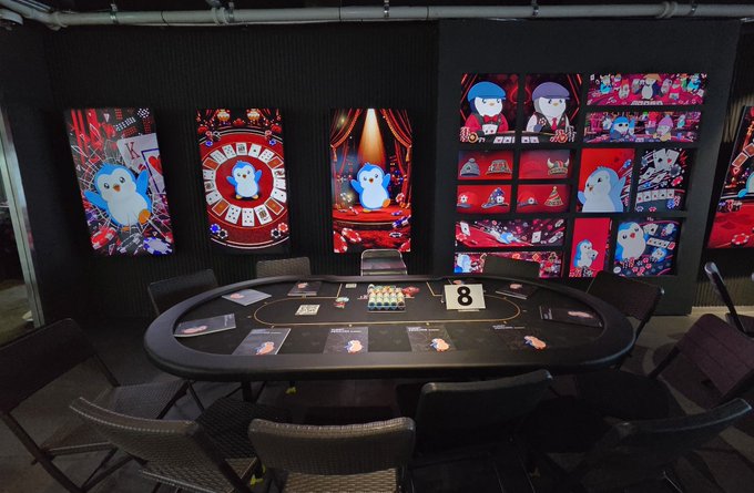 An oval gaming table marked with the number "8" sits surrounded by chairs in a room decorated with vibrant Pudgy Penguins artwork, including a roulette wheel, stage, and character displays. The space reflects the post's excitement about spending the day with @pudgypenguins, with the cartoon penguins and gaming setup creating a fun, themed experience.