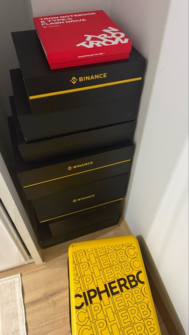 A stack of Binance-branded boxes sits in a corner next to a yellow CipherBC merchandise bag and a red Tron Notebook "Flash Drive" package, showcasing part of the crypto collectibles and merch collection that includes items from exchanges like OKX and Mexc.