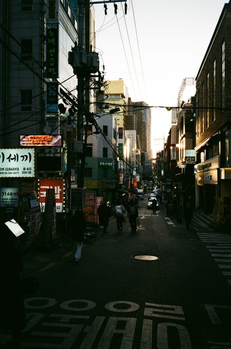 A dimly lit Japanese street scene at dusk, lined with storefronts featuring illuminated signs, overhead power lines, and pedestrians walking down the road.