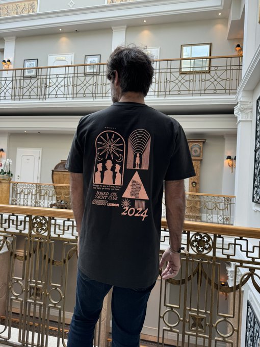 A man stands facing away from the camera on an ornate hotel balcony, wearing a black t-shirt featuring the "Bored Ape Yacht Club" logo and the year "2024" on the back. The shirt's design, which includes an ape silhouette and rocket imagery, visually represents his pride as a member of the exclusive NFT community described in the caption.