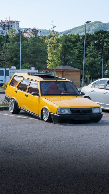 A modified yellow station wagon with lowered suspension and custom wheels parked in a lot, featuring red interior seats and a black hood.
