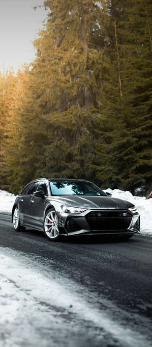 A dark gray Audi RS7 sedan parked on a snow-covered road with evergreen trees in the background, illuminated by warm sunset light.