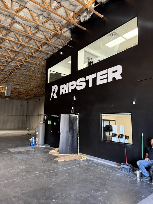 An interior shot of a building under construction showing the 'RIPSTER' logo in white on a black wall, with exposed wooden roof trusses and a worker sitting nearby.