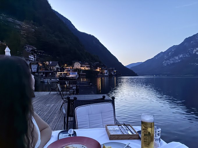 A serene evening view from a lakeside restaurant deck, showing a woman seated at a table with a drink while looking out over the water towards a village nestled between mountains.