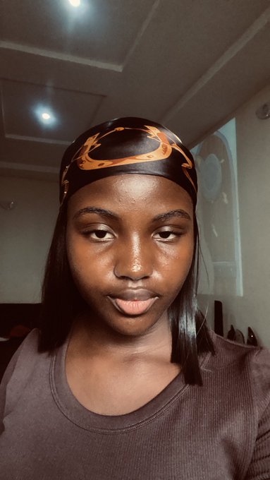 A young woman wearing a black and orange patterned headband and a brown shirt poses for a selfie in an indoor setting.