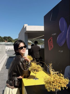 A woman wearing sunglasses and a dark top makes a peace sign while posing outdoors near a modern building with sculptural artwork and decorative plantings.