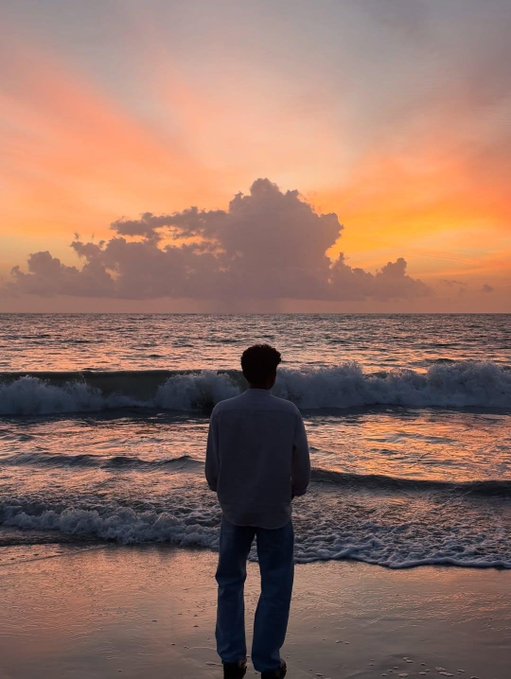 A person stands on a sandy beach gazing at a vibrant orange and pink sunset over ocean waves with dramatic cloud formations.