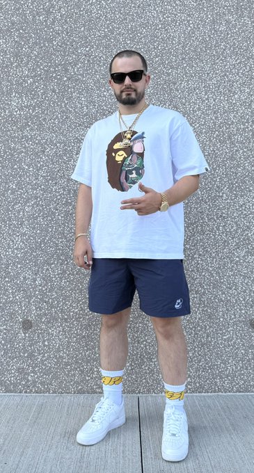 A man wearing sunglasses, a graphic t-shirt, navy shorts, and yellow-patterned socks poses for a full-body photo against a gray textured wall.