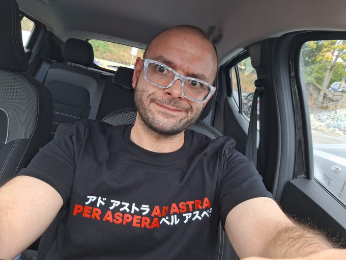 A bald man with glasses taking a selfie in a car, wearing a black t-shirt with red and white text reading 'アド アストラ PER ASPERA アストラ'.
