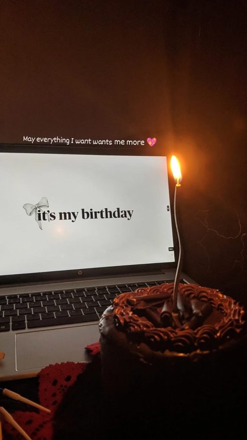 A laptop displays the text "it's my birthday" with a bow graphic, alongside a lit candle in a chocolate birthday cake. The cozy scene captures the personal birthday celebration mentioned in the post, with the caption "May everything I want wants me more 💕" adding a hopeful tone.