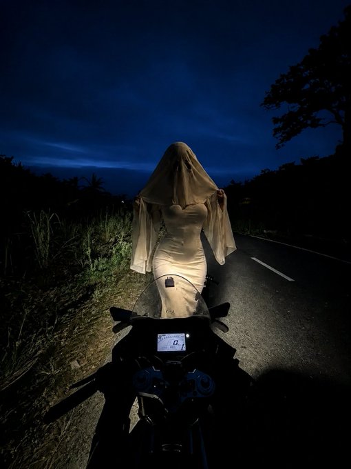 A nighttime scene showing a person in a white dress with a veil standing on a road, viewed from the perspective of a motorcycle rider.