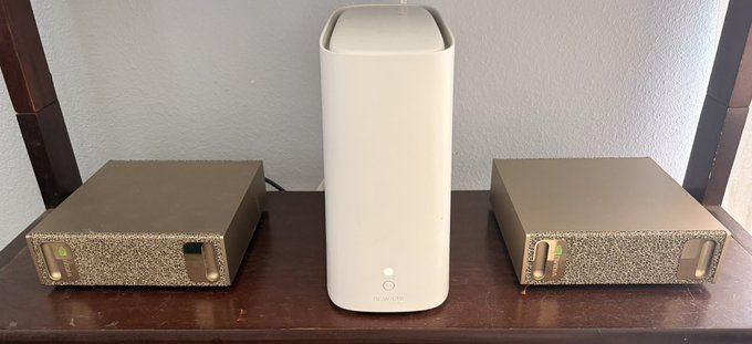 A Linksys Velop mesh Wi-Fi node stands in the center on a wooden shelf, flanked by two textured bronze-colored external hard drives with green status lights.