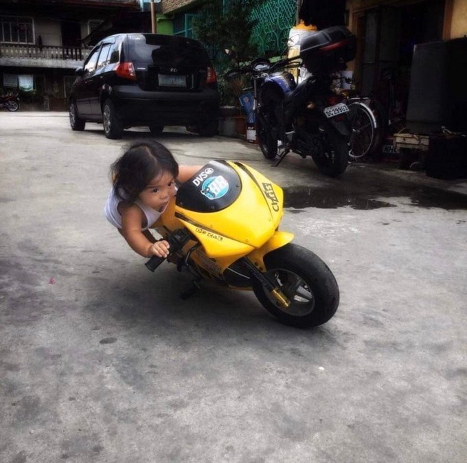 A young child playing with a yellow toy motorcycle on a concrete driveway, with a black car and motorcycles parked in the background.
